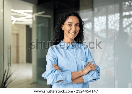 Portrait of happy and successful business woman, boss in shirt smiling and looking at camera inside office with crossed arms, Hispanic woman with curly hair in corridor.