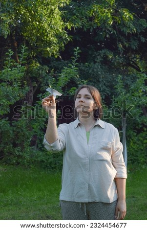 Balance concept. Front view happy adult woman launches paper airplane in bright sun rays in beautiful landscape garden at sunset. Serene female person uses origami to express harmony state