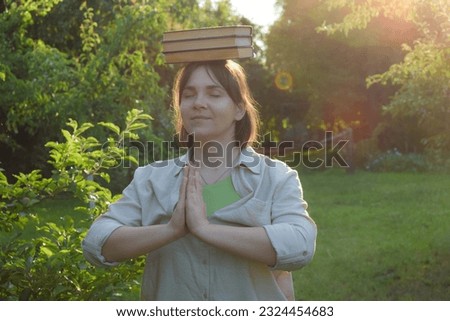 Young adult woman balancing books on head with closed eyes outdoors at sunset. Balance concept, life work equilibrium analogy. Golden hour sun rays at green landscape garden park on background