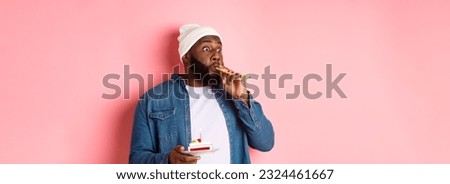 Happy Black hipster celebrating birthday, blowing party whistle, holding bday cake with candle, standing over pink background.