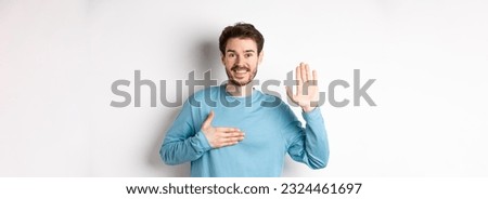 Smiling hosent man raising arm and hold hand on heart, making promise tell truth, swear or give oath, standing over white background. Royalty-Free Stock Photo #2324461697