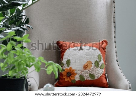 Looking past a potted basil plant to a beautiful, decorative pillow with a pumpkin, bird, flowers, and leaves embroidered onto it.