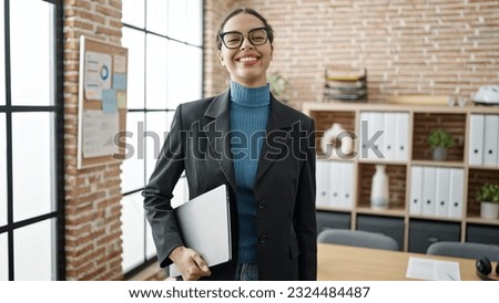 Young beautiful hispanic woman business worker smiling confident holding laptop at office
