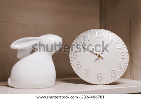 white clock and figurine of white cute porcelain rabbit stand on the shelf, home decor