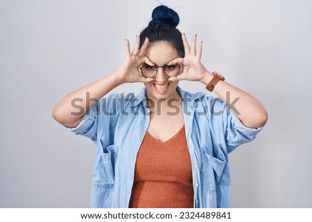 Young modern girl with blue hair standing over white background doing ok gesture like binoculars sticking tongue out, eyes looking through fingers. crazy expression. 