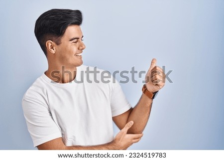 Hispanic man standing over blue background looking proud, smiling doing thumbs up gesture to the side 