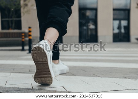 Legs of a girl in White Leather Sneakers Crosses the Street on a Pedestrian Crossing, close-up rear view. Summer Shoes, Pedestrian.