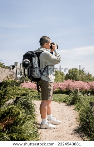 Side view of short haired and young female travel photographer taking photo on digital camera and standing on scenic nature at background, travel photographer, trekking through landscape, summer