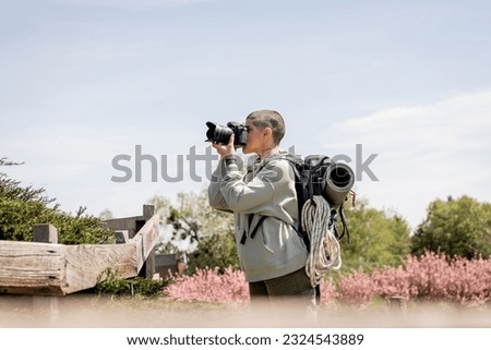 Side view of young short haired and tattooed hiker with backpack taking photo on digital camera while standing near wooden fence and scenic landscape, travel photographer