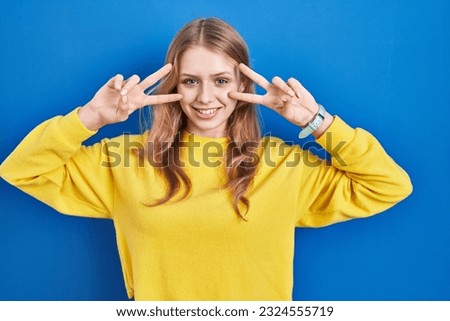 Young caucasian woman standing over blue background doing peace symbol with fingers over face, smiling cheerful showing victory 