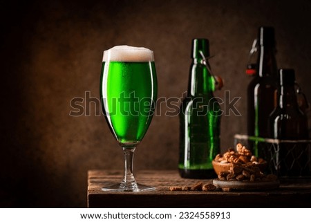 Glass of green beer with foam head on dark wooden background with empty bottles and beer snacks. St. Patricks Day concept. Copy space.
