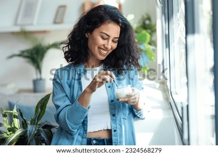 Shot of happy beautiful woman eating yogurt while standing in living room at home. Royalty-Free Stock Photo #2324568279