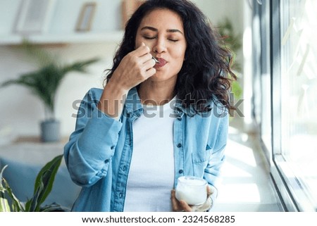 Shot of happy beautiful woman eating yogurt while standing in living room at home. Royalty-Free Stock Photo #2324568285