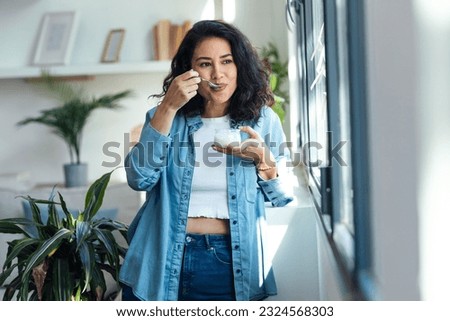 Shot of happy beautiful woman eating yogurt while standing in living room at home. Royalty-Free Stock Photo #2324568303