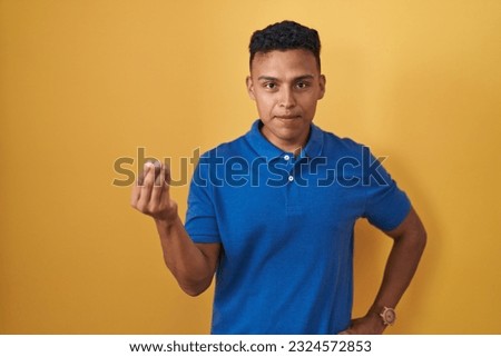 Young hispanic man standing over yellow background doing italian gesture with hand and fingers confident expression 