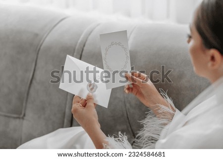 Portrait of a beautiful girl bride with a stylish hairstyle and makeup in a white robe, sitting on a sofa and holding a wedding invitation. Free space