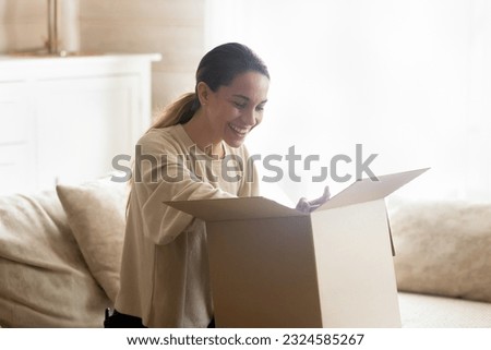 Let us look what is inside. Curious happy millennial woman client customer shopper buyer sitting on sofa at home opening unboxing cardboard box with mail package glad to get delivery easy and quick Royalty-Free Stock Photo #2324585267