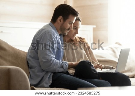 Using laptop. Concentrated young husband and wife sitting together on comfy sofa using laptop computer, making payments or purchases online, browsing internet sites, reading posts at social networks Royalty-Free Stock Photo #2324585275