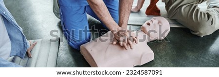 first aid seminar, hands-on learning, cropped view of paramedic showing chest compressions on CPR manikin near young participants in training room, life-saving skills and techniques concept, banner Royalty-Free Stock Photo #2324587091