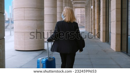 Sttylish female manager in safety mask with suitcase walking outside office building. Rear view of mature businesswoman in protective mask with luggage walk outdoors
