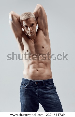 Portrait of handsome young man with blonde hair, musuclar fit body posing shirtless in jeans against grey studio background. Concept of male natural beauty, body care, health, sport, fashion, ad Royalty-Free Stock Photo #2324614739