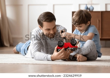 Loving young Caucasian father sit on floor feel playful repair fix toy car together with little 8s son. Caring happy dad play with automobiles with small preschooler boy child at home on weekend. Royalty-Free Stock Photo #2324629919