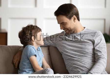 Happy young father and little son sit on couch in living room talk chat on leisure family weekend. Smiling loving Caucasian dad and small boy kid child have conversation, share secrets at home. Royalty-Free Stock Photo #2324629921
