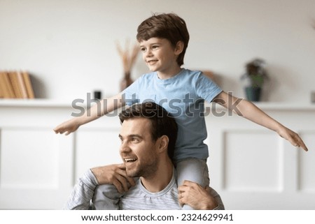 Excited small Caucasian 7s boy child sit on young father shoulder back have fun at home on family free weekend together. Happy little son kid play with dad, engaged in funny game activity. Royalty-Free Stock Photo #2324629941