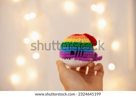 Hand holding rainbow crochet handmade plush shark on white background with lights bokeh. Pride symbol