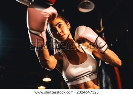 Asian female Muay Thai boxer punching in fierce boxing training session, delivering strike to her sparring trainer wearing punching mitts, showcasing Muay Thai boxing technique and skill. Impetus Royalty-Free Stock Photo #2324647325
