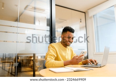 Smiling young indian businessman using phone typing messages and browsing, man in casual wear sitting in front of laptop in office, holding smartphone, male student chatting online