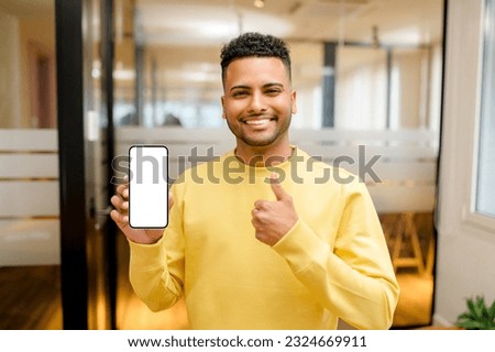 Cheerful handsome male office employee holding and showing smartphone in contemporary coworking space, smiling arab student showing empty blank phone screen and thumb up in library, recommending app