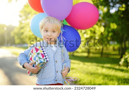 Child going to congratulate a friend on his birthday. Toddler holding bundle of colorful balloons and gift in a festive box. Cute little boy celebrate party with colorful balloons.