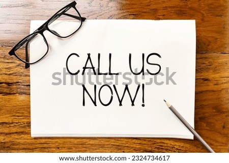 Call us now handwriting text on blank notebook paper on wooden table with pencil and glasses aside. Business concept to inform customer to make a phone call.