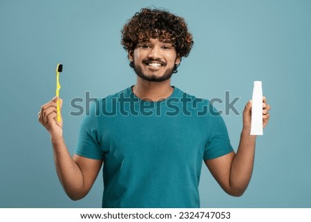 Handsome smiling Indian man looking in mirror, holding toothbrush and tube of toothpaste looking at camera isolated on blue background. Concept of dental care and health