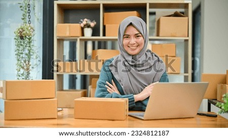 A portrait of a successful young Asian Muslim female online seller sits at her working desk with a laptop and cardboard boxes on a table.
