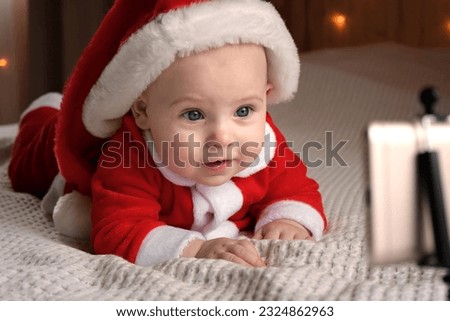 Smiling Baby Wearing Red Santa Claus Costume Making Video Call looking at Phone. Christmas Online Congratulations. Cute Child Communicates and Has Fun, Using Smartphone. Merry Xmas and Happy New Year.