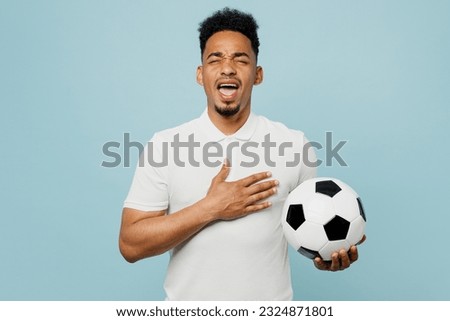 Young man fan wearing basic t-shirt sing national country anthem cheer up support football sport team holding in hand soccer ball watch tv live stream isolated on plain blue color background studio Royalty-Free Stock Photo #2324871801