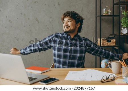 Successful happy minded cheerful employee business Indian man he wearing casual blue checkered shirt do hands stretching look aside rest relax sit work at office desk with laptop pc computer indoors Royalty-Free Stock Photo #2324877167