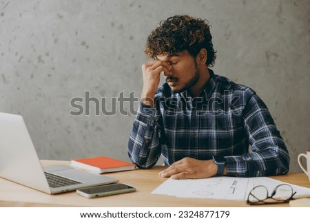 Troubled sad preoccupied tired employee business Indian man he wearing casual blue checkered shirt keep eyes closed rub put hand on nose worry sit work at office desk with laptop pc computer indoors