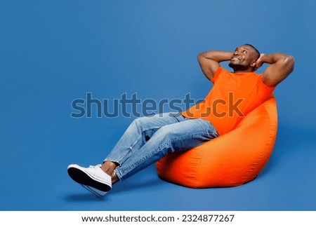Full body young man of African American ethnicity he wear orange t-shirt sit in bag chair hold hands behind neck isolated on plain dark royal navy blue background studio portrait. Lifestyle concept