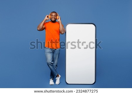 Full body young man of African American ethnicity wear orange t-shirt headphones big huge blank screen mobile cell phone smartphone with area listen music isolated on plain dark navy blue background
