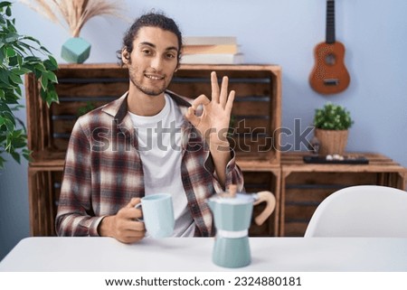 Young hispanic man drinking coffee from french coffee maker doing ok sign with fingers, smiling friendly gesturing excellent symbol 