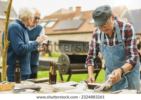 Senior Friends Preparing For Barbecue Party - A senior man with a hat prepares barbecue food as his friends, also seniors, chat. One holds a harmonica. Beers and board games adorn the table.
