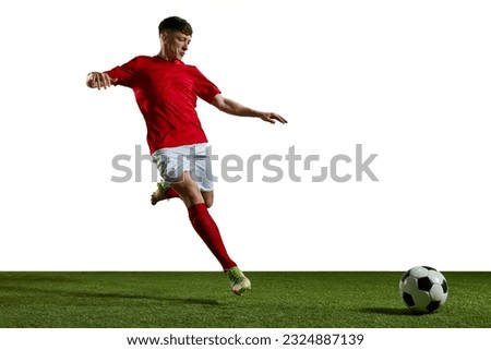 Young man in red uniform, football player in motion, playing, kicking ball on sports field against white background. Concept of professional sport, action, lifestyle, competition, hobby, training, ad Royalty-Free Stock Photo #2324887139