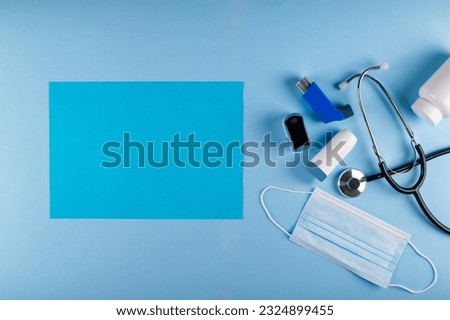 Medical mask, stethoscope, oximeter, bottle of pills and inhalers on light blue background with vibrant blue rectangular sheet of paper for text placement. Asthmatic breathing symptoms Concept Royalty-Free Stock Photo #2324899455