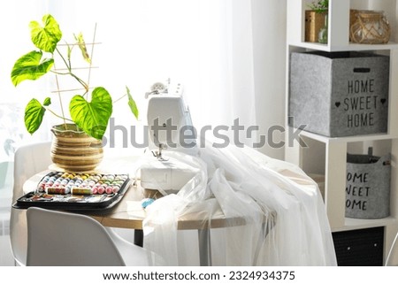 Sewing machine on a round table in the white interior of the house near the window with a transparent curtain, and a home plant in a pot - a modern interior. hobby, home business
