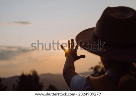 Silhouette of dreaming little girl in hat pulls hand to warm sun. Religion helping hand. Preteen child enjoy beautiful summer nature during amazing sunset or sunrise. Prayer in religion concept Royalty-Free Stock Photo #2324939279