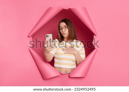 Cute attractive brunette woman wearing striped t shirt posing in torn pink paper wall holding smart phone making selfie or having video call sending air kissing.