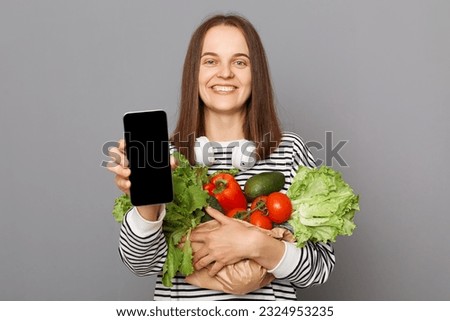 Cheerful woman embracing fresh vegetables returns from supermarket wearing striped  shirt isolated over gray background showing mobile phone empty display.